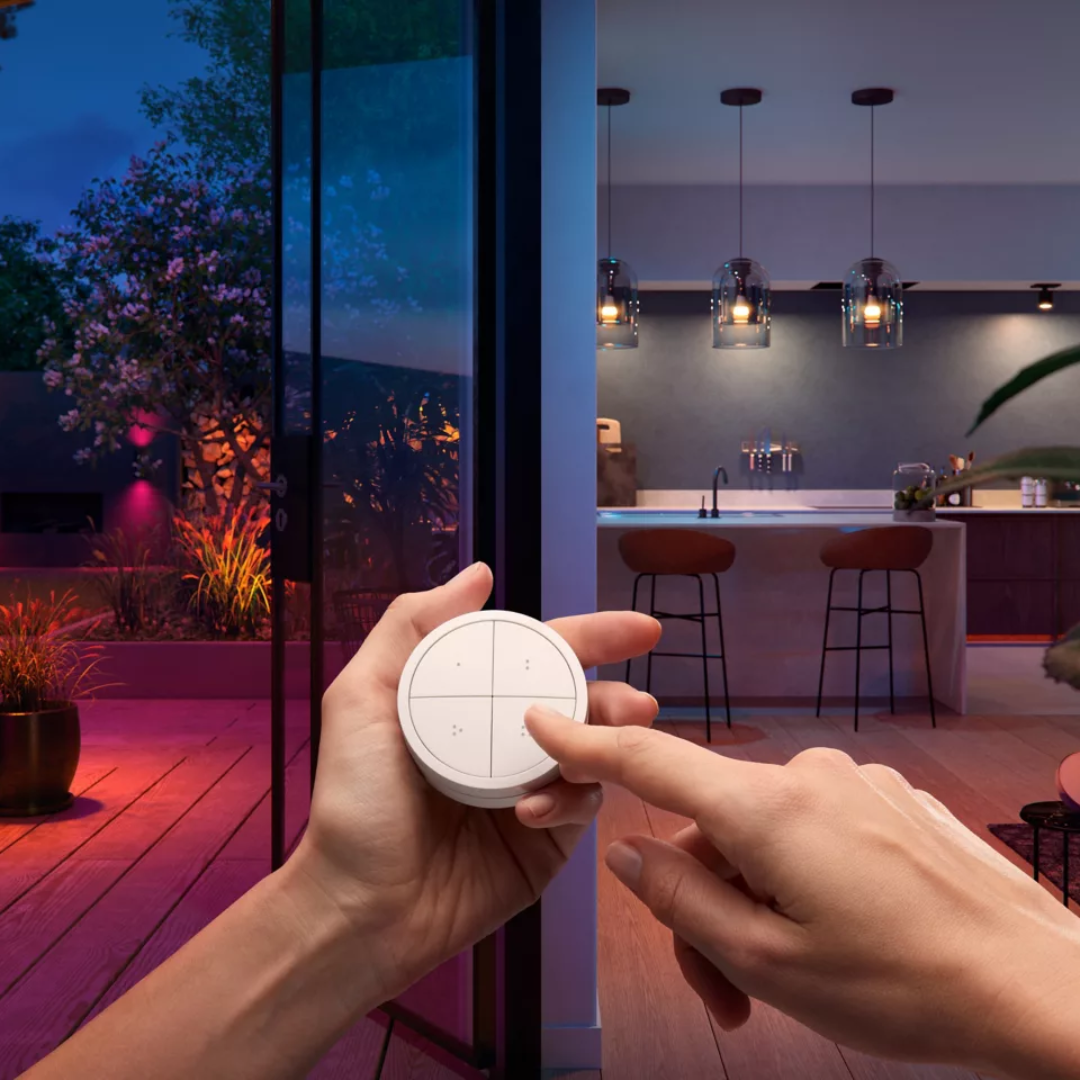 Ovladač Philips Hue Tap Dial. Ruka drží kulatý bílý ovladač, který je bezdrátový a umožňuje plynulé stmívání a přepínání scén v pozadí.