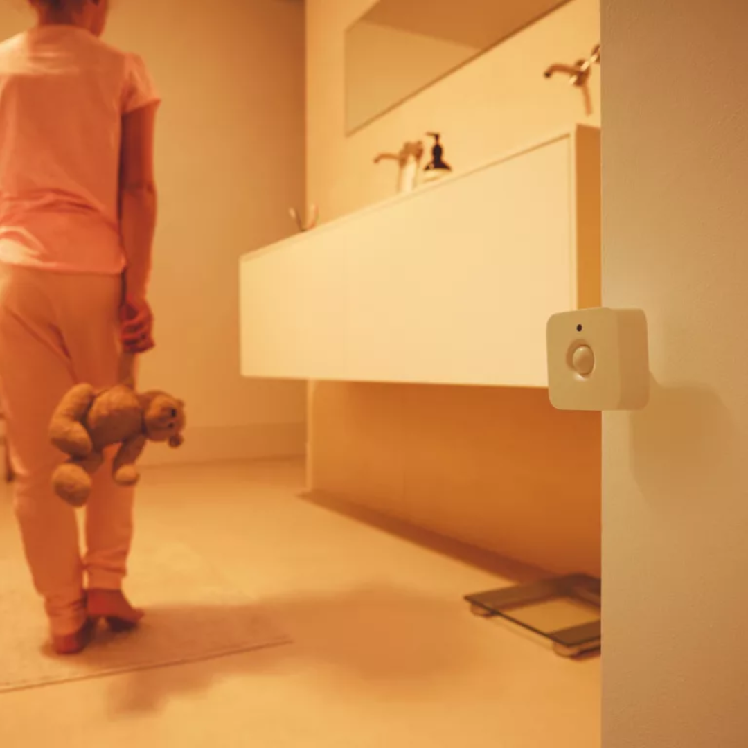 Vnitřní pohybový senzor Philips Hue v koupelně. Osoba s plyšovým medvědem prochází kolem senzoru na stěně, který automaticky rozsvítí teplé světlo.